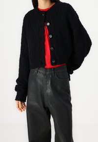 Cardigan noir en tricot avec un motif texturé, col rond et cinq gros boutons. Porté sur une chemise rouge et associé à un pantalon noir.