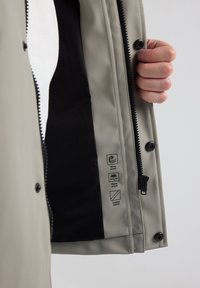 Waterproof jacket with a beige outer layer. Features a black liner, front zipper, and four icons indicating windproof and waterproof qualities.
