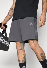 Mand iført grå Adidas shorts, sorte Adidas sokker og holder en sort Adidas taske, med flere farverige tatoveringer på ben og arme.