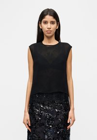 Femme portant un haut noir transparent sans manches sur un soutien-gorge noir et une jupe noire à sequins, debout devant un fond blanc uni.