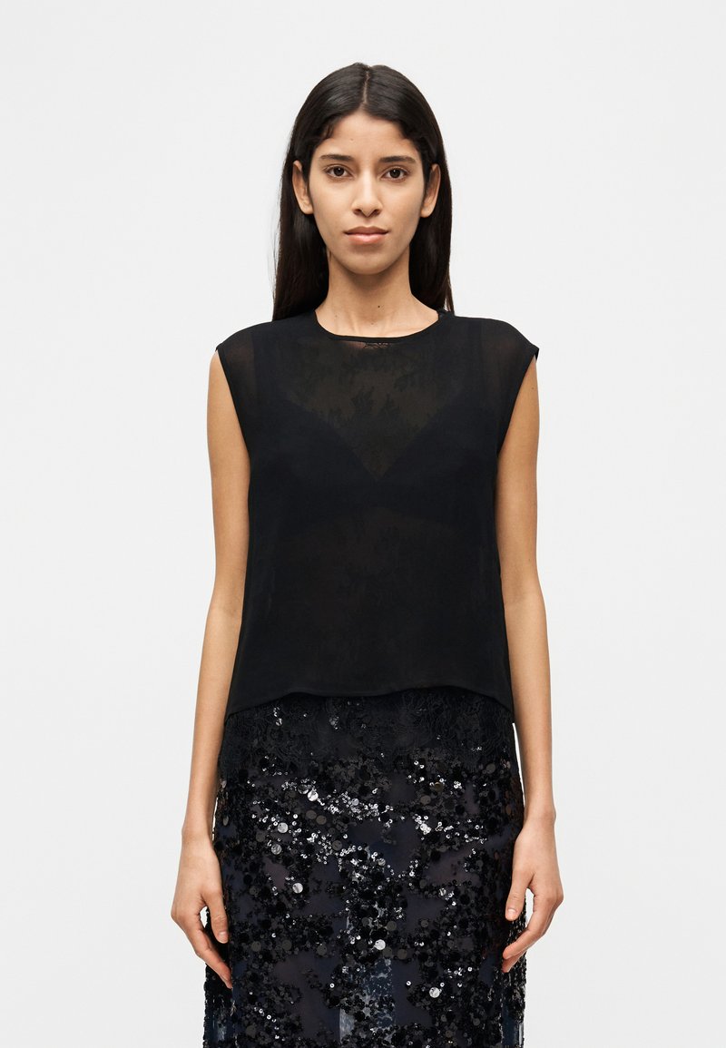 Femme portant un haut noir transparent sans manches sur un soutien-gorge noir et une jupe noire à sequins, debout devant un fond blanc uni.