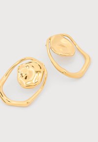 Ασύμμετρα σκουλαρίκια σε χρυσό τόνο με αφηρημένα, ακανόνιστα κυκλικά σχήματα και υφή σφυρήλατου μετάλλου σε λευκό φόντο.