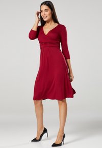 Bordeauxfarbenes knielanges Kleid mit V-Ausschnitt und Dreiviertelärmeln. Hergestellt aus glattem Stoff mit tailliertem Oberteil und schwingendem Rock. Schwarze High Heels.