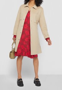 Un manteau en laine beige avec deux poches avant, des boutons argentés et un col classique, associé à une robe florale rouge et des mocassins noirs à semelles épaisses.