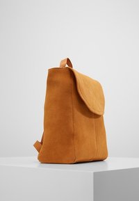 Mochila de ante color beige con solapa plana, forma cuadrada y un asa superior única. Presenta una textura suave y un diseño minimalista.