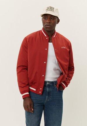 Veste bomber rouge avec des rayures blanches sur le col et les poignets, avec des boutons-pression, portée sur un t-shirt blanc et un jean bleu.