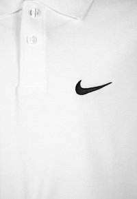 Valkoinen poolopaita, valmistettu struktuurisesta kankaasta, jossa on kolmen napin aukko ja vasemmassa rinnassa musta kirjailtu Nike-logo.