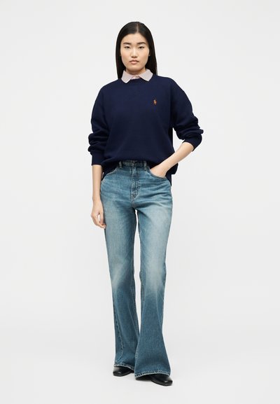 Νεαρή γυναίκα στέκεται με το χέρι στην τσέπη φορώντας μπλε σκούρο φούτερ πάνω από παστέλ ροζ πουκάμισο με γιακά, ανοιχτό μπλε τζιν καμπάνα και μαύρα παπούτσια.