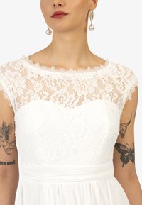 Witte jurk met kanten lijfje met bloemmotieven, korte mouwen en een getailleerde taille; gecombineerd met gouden ringoorbellen en parelaccenten.