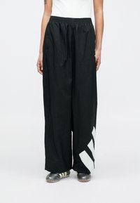 Schwarze Hosen mit weitem Bein und elastischem Bündchen. Versehen mit einem weißen diagonalen Streifenmuster auf der unteren linken Seite. Hergestellt aus leichtem Material.