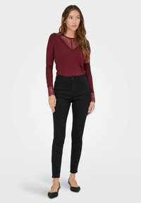 Bordeaux lange mouw top met kantaccenten bij de halslijn en manchetten, gecombineerd met zwarte skinny broek en zwarte textuur flatjes.