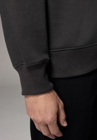 Ciemnoszary sweatshirt z prążkowanym mankietem, o gładkiej fakturze i prostym designie, prezentowany obok ręki opierającej się o materiał.