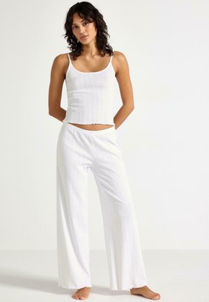 Kvinde stående barfodet iført hvid ribbet tanktop og vide bukser mod en ensfarvet hvid baggrund.