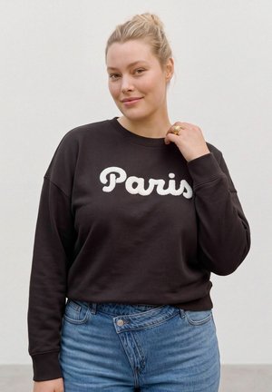Une femme souriante porte un sweat foncé avec l’inscription « Paris » et un jean bleu. Elle tient le col devant un fond uni et clair.