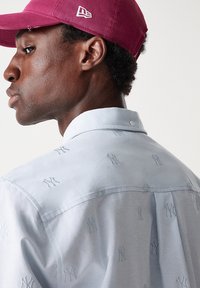 Camicia azzurro chiaro con logo ricamato discreto dei New York Yankees, dotata di colletto standard e tessuto leggero. Cappellino bordeaux sopra.