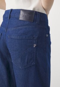 Gros plan sur un jean en denim bleu foncé avec une poche arrière comportant un petit logo métallique et un patch en cuir portant la marque Dondup.