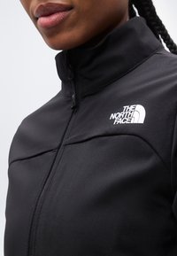 Κοντινό πλάνο ενός ατόμου που φοράει μαύρο μπουφάν με φερμουάρ της The North Face στο στήθος και έχει ορατά πλεξούδες στα μαλλιά.