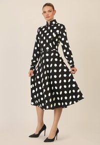 Schwarzes Midi-Kleid mit weißen Punkten, langen Ärmeln, einem Kragen und einer taillierten Taille, die nach unten ausgestellt ist. Dazu schwarze High Heels.