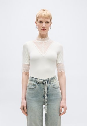 Rövid szőke hajú nő, fehér csipkés szegélyű, fél ujjú felsőt és világoskék, magas derekú farmert visel, amelyhez megkötős öv tartozik, előre néz.