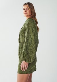 Olivgrünes Kleid mit einer gewellten Textur, langen Ärmeln, tailliertem Bund und einer Länge über dem Knie. Schlichtes, elegantes Design ohne sichtbare Muster.