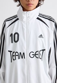 Valkoinen adidas-takki, jossa on mustat raidat, "TEAM GEIST" -teksti ja numero "10" etupuolella. Korkea kaulus ja vetoketjukiinnitys.
