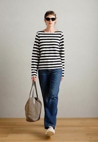 En svart og hvit stripete langermet topp kombinert med blå jeans; modellen holder en grå totebag; hun har på seg svarte solbriller og hvite joggesko.
