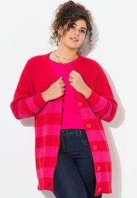 Cardigan rouge tricoté avec des manches rayées roses, doté de boutons ronds, porté sur un haut rose et un jean foncé. Texture lisse, coupe décontractée.