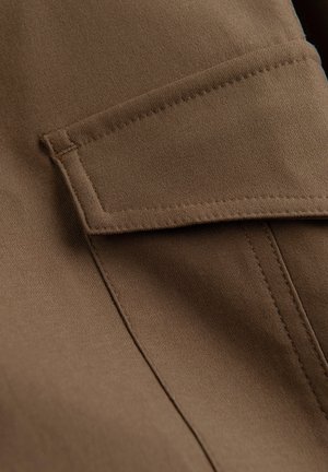 Brun stof tæt på viser en rektangulær klap lomme med syningsdetaljer på glat tekstil materiale.