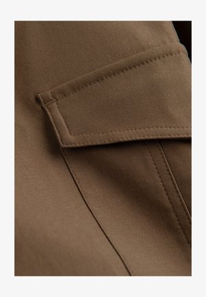 Brun stof tæt på viser en rektangulær klap lomme med syningsdetaljer på glat tekstil materiale.