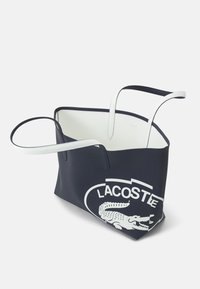 Sac cabas bleu marine avec poignées et intérieur blancs, présentant un grand logo Lacoste blanc avec un dessin de crocodile d'un côté.