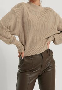 Beżowy sweter dzianinowy z wysokim dekoltem i żebrowaną teksturą, w zestawieniu z ciemnobrunatnymi skórzanymi spodniami z przednimi kieszeniami.