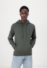 Hoodie vert foncé avec une poche avant, capuche avec cordon de serrage ajustable, poignets côtelés et un petit logo noir sur la poitrine.