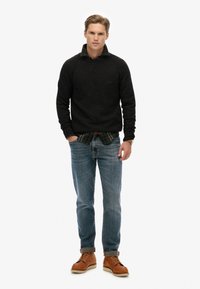 Zwarte trui, groen geruit shirt eronder, blauwe jeans, bruin leren laarzen. Casual outfit met een ontspannen pasvorm en omgeslagen mouwen op de jeans.