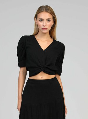 Femme aux longs cheveux blonds portant un crop top noir à manches courtes, col en V et devant torsadé, et une jupe noire taille haute sur fond gris.