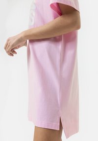 T-shirt en coton rose avec des manches courtes et des fentes sur les côtés. Le tissu a une texture lisse et il y a un discret design graphique blanc à l'avant.