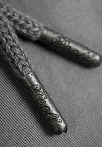 Cordones de zapatos trenzados grises con agletes metálicos que presentan letras en relieve, descansando sobre un fondo de tela gris texturizada.