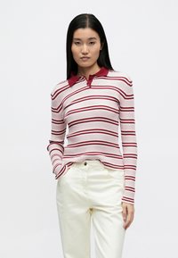 Csíkos, hosszú ujjú rózsaszín-piros póló ing bordázott textúrával, piros gallérral és karcsúsított szabással, fehér nadrággal párosítva.