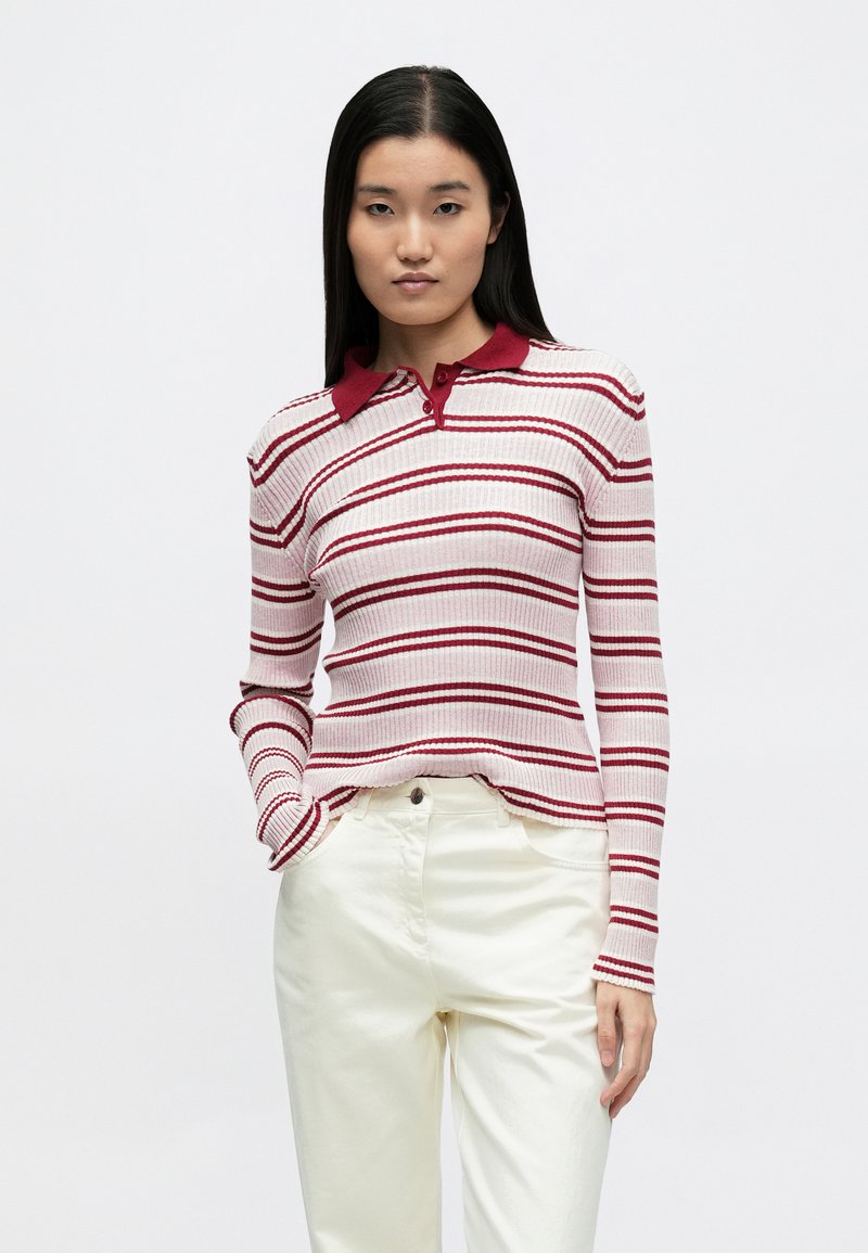 Csíkos, hosszú ujjú rózsaszín-piros póló ing bordázott textúrával, piros gallérral és karcsúsított szabással, fehér nadrággal párosítva.