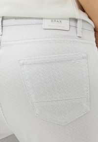 Pantalones vaqueros rayados de gris claro que presentan un bolsillo trasero y una etiqueta de cuero blanco con el logo "BRAX" grabado. La textura es suave, con líneas verticales sutiles.