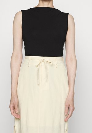 Mouwloze zwarte top gecombineerd met een crèmekleurige rok met hoge taille, gestrikt met een strik in de taille, gedragen door een staand persoon tegen een effen achtergrond.