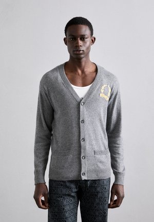 Grå cardigan med v-hals og knapper, med ribbede manchetter og kant. Broderet gul detalje på venstre bryst. To forlommer.