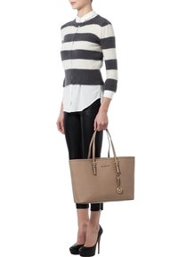 Beige schoudertas met dubbele hengsels, met een gladde lederen textuur, gouden hardware en een charm. Het model draagt een gestreept sweater en zwarte broek.