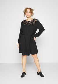 Vestido preto com mangas longas, painel de renda na gola, cintura cingida com cinto e saia em A. Tecido suave com uma textura leve.