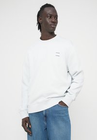 Ανοιχτό γκρι φούτερ από απαλό ύφασμα, με ριμπ μανσέτες και τελείωμα. Μικρή μπλε λεπτομέρεια με το λογότυπο "SAMSØE" στο μπροστινό μέρος αριστερά στο στήθος. Φορεμένο με μπλε τζιν.