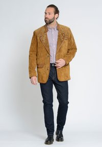Bruine suede jas met een verfijnde snit, voorzien van geborduurde accenten en contrasterende leren afwerking. Gekleed met donkere jeans en zwarte laarzen.