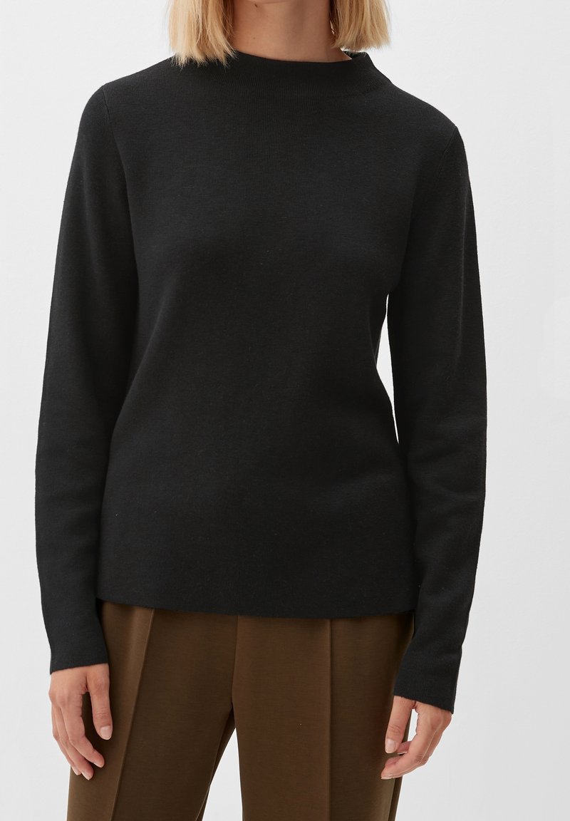 Pull noir à manches longues avec col montant, fabriqué dans un tissu doux et texturé. La coupe est décontractée et se combine avec un pantalon marron.