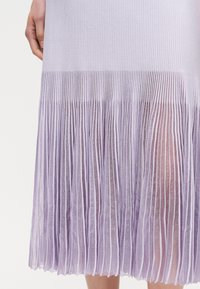 Λεβάντα πλισέ φούστα με διαφανές ύφασμα επικάλυψης, που δείχνει διακριτικές κάθετες ρίγες και μερική θέα σε πόδια και ένα χέρι που ακουμπά δίπλα της.