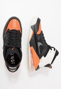 Černé a oranžové sportovní tenisky s síťovinovými panely, perforovanou špičkou, bílými akcenty a viditelnou vzduchovou jednotkou v patě.