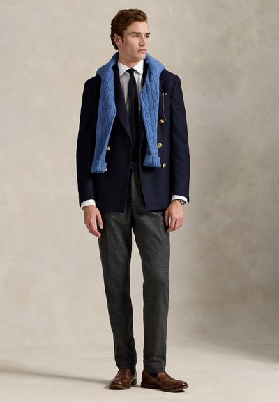 Mand iført marineblå dobbelttradet blazer med guldknapper, hvid skjorte, sort slips, grå bukser, brune sko og blå sweater draperet over skuldrene.