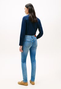 Γυναίκα με μακριά σκούρα μαλλιά, που φοράει μπλε σκούρο πουκάμισο μακρυμάνικο, ανοιχτό μπλε τζιν και ταμπά μοκασίνια, όρθια με την πλάτη προς τα εμπρός.